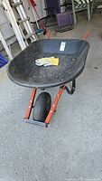 View of entire wheelbarrow with black PVC tray and orange handles, work gloves placed inside tray on concrete floor.