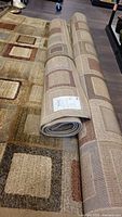 Two rolled area rugs and partially unrolled largest rug showing matching square and rectangle pattern in earth tone colors