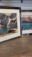 Photo of two framed art prints leaning against a wall, showing landscape scenes of mountains and lakes with visible white snow patches, wrapped in protective plastic.