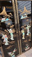 Two black lacquer panels with detailed 3D stone inlays of traditional Asian figures and mountain landscapes with gold accents.