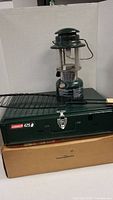 Coleman 425 two-burner stove stacked on original box with green Coleman Easi-Lite lantern on top and fish holder rack visible.