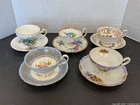 Five assorted porcelain cups and matching saucers with floral patterns and gold accents shown together on a black surface.