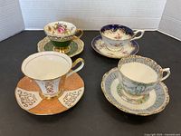 All four porcelain cups and saucers displayed on black surface, showing distinct patterns and gold accents.