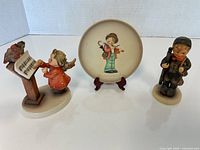 Two Hummel figurines and a small plate displayed on a white surface.