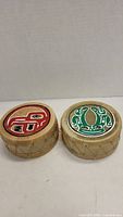 Pair of small round indigenous drums side by side showing top painted animal designs in red and green.