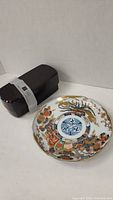 Photo showing black Japanese lunch box sealed with a white band next to Japanese Imari decorative plate