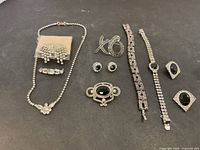 All items of costume jewelry are displayed on a dark background, showing necklaces, earrings, brooches, bracelets, and rings primarily with clear rhinestones and black stones.