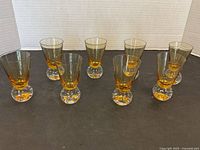 Eight amber tinted cocktail or cordial glasses arranged in a row on a black surface, showing clear ball footed bases.