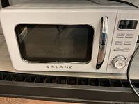 Front view of the Galanz white microwave oven showing the door with a chrome handle, digital control buttons, and dial timer.