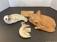 Lot showing three figurines: a white seal lying on its side, a brown deer lying curled up, and a Snowbaby baby on a crescent moon with pink bird figurine. Included is a small printed card.