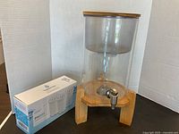 Clear water container approximately 12 inches tall with wooden lid and wooden stand. Box with replacement filters visible next to it.