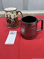 Two beer steins placed side by side on a red surface, each with distinct materials and finish. One is ceramic with detailed artwork and the other is silver-tone metal.