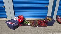 Wide photo showing two bins of Christmas ornaments, various garlands, and Christmas linens laid out outdoors on a mat in front of a blue storage unit door.