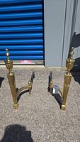 Pair of brass fireplace andirons outside near a blue storage unit door, showing overall size and shape.