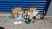 Wide shot showing various sizes of picture frames, a lamp, Wedgwood saucers, and other small decor pieces arranged on pavement against a blue corrugated metal wall
