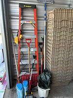 Full view of orange fiberglass step ladder, snow shovels, garden tools, white bucket with green tarp, and beige storage bins