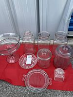 Lot displayed on red cloth with glassware including trifle bowl, small plate, platter, and five glass canisters with lids arranged in two rows.