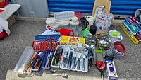 Wide shot of assorted kitchenware including cutlery sets, trays, containers, French press, and various small appliances and utensils arranged on a ground surface.