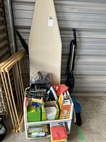 Laundry and cleaning lot showing ironing board, drying rack, various cleaning products, sewing kit inside bin, and vacuums leaning on the wall.