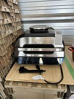 Front view of Ninja Foodi Smart XL Grill showing stainless steel construction and included cleaning brush and power plug in front.