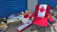 Wide view of picnic lot showing two red folding chairs with large Canadian maple leaf design, picnic baskets, plates, cups, and other picnic accessories arranged on surfaces and ground.