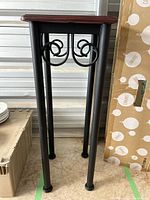 Full view of the plant stand showing tubular metal legs with curled metal supports and square wood top with a glass pane in the center.