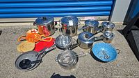 Wide view of all pots, pans, trivets, colanders and bowl arranged outside on asphalt surface.