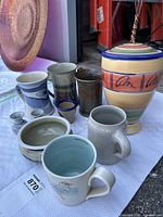 Photo showing the entire pottery lot including tall lidded vase, mugs, small vases, small cup, and round bowl arranged on a table