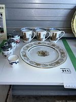 Photo displaying the set of three teacups with architectural royal prints, Aynsley decorative plate with gold filigree border and coat of arms, three Royal Worcester egg coddlers, and Adderley shot glass