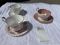 Photo showing three vintage teacups with matching saucers arranged on a white quilted surface, displaying floral patterns and gold trim.