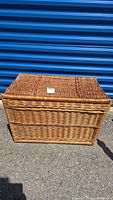 Front view of large rectangular wicker basket with hinged lid closed, showing woven wicker material and a small tag on the lid.