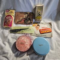 Photo showing multiple vintage beauty products including Avon lipstick box, boxed Meta product, nylon hair nets, hairdresser comb, and two compact powder containers.