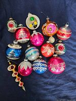 Photo shows 14 vintage Christmas ornaments on dark blue fabric background including various round glass baubles, one figurine ornament, and one icicle shape ornament