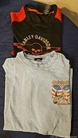 Two folded shirts. One black with orange accents and skull logo, one light blue with motorcycle and flag graphic.