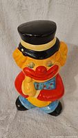 Front view of the vintage chalkware clown bank showing the clown with a black top hat, yellow hair, red jacket, blue polka dot bow tie, and blue pants.