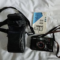 Canon camera with lens, autofocus and flash visible, on white surface next to black leather case and folded Canon Z35/Z85 manual.