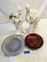 Full shot showing two white ceramic Asian-style figurines and two bowls, one blue ceramic and one red lacquer with gold bamboo design
