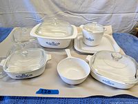 Five Corning Ware Blue Cornflower stoneware pieces including casseroles with clear glass lids and one plain white microwave tray, all arranged on a beige mat over blue cloth. Lids and casseroles in good condition with some scuffing and marked chips on lids.