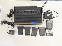 Photo showing entire Atari 2600 console, joystick controller, power supply, and 6 game cartridges laid out flat. Console shows some wear and has a blue sticker labeled '225'.