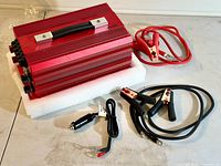 Red 2000 watt power inverter with handle, resting on white foam, alongside multiple connecting cables including battery clamps and cigarette lighter plug.