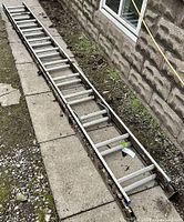 Full view of aluminum extension ladder laid out straight on concrete and soil ground, showing its length and condition.