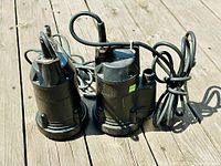 Two black submersible sump pumps placed side by side on wooden decking with cords and fittings visible.