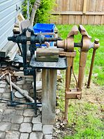 Two post vises clamped side by side to a wooden support, one painted primer brownish, the other dark blue/black; support legs extend to ground