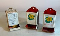 Three vintage tin litho match holders displayed front side showing advertising and floral designs.