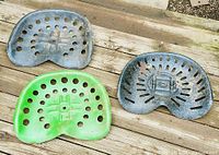 Three cast iron tractor seats arranged on wood surface, one green and two unpainted with rust and wear