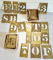 Brass stencils laid out on a surface, including letters E, S, T, F, O, and numbers 0-9, with two small containers holding additional stencils