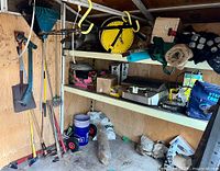 Wide shot of the shed wall showing multiple garden tools hanging on the wall, shelves with the BBQ grill, storage boxes and bags, and various gardening items including a wheelbarrow and hose reel