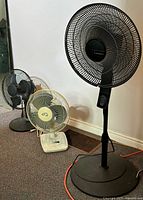 View of all three fans together: one black Lasko floor fan and two smaller table fans (one black, one off-white/yellow)
