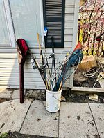 Photo showing a 5-gallon bucket filled with multiple fishing rods and a fishing net leaning against the side of a house.