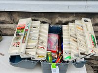 Top view of open tackle box showing multiple fold-out trays with assorted fishing lures, hooks, packaged fishing accessories, and soft plastics